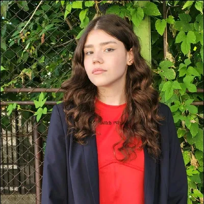 Ana Farazică, wearing a red top and dark jacket, standing in front of green leaves.