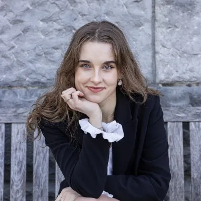 Kinga Marchlewska, resting her chin on her hand, wearing a dark blazer over a white ruffled blouse, seated on a wooden bench in front of a stone wall.