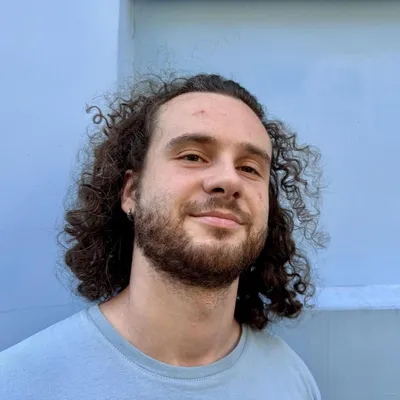 Maciej Czerniak, smiling, with shoulder-length curly hair and a short beard, wearing a pale blue t-shirt, in front of a light blue-grey wall.